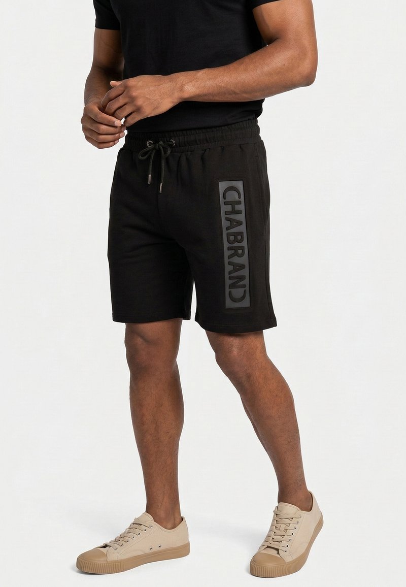 Hombre vistiendo pantalones cortos negros con cordón y el logo "CHABRAND" en la pierna izquierda, y zapatillas beige, de pie contra un fondo blanco liso.