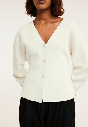 Femme portant un cardigan blanc à col en V avec trois boutons et manches longues, associé à un pantalon noir ample sur un fond uni.
