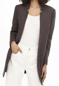 Blazer largo gris hecho a medida con cuello de solapa, confeccionado en tela suave; combinado con una camiseta sin mangas blanca y pantalones de color claro.