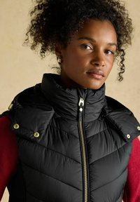 Czarna kamizelka puchowa z wysokim kołnierzem, złotym zamkiem błyskawicznym i zatrzaskami. Posiada pikowany wzór i miękką fakturę. Nosi się ją na czerwonym swetrze.
