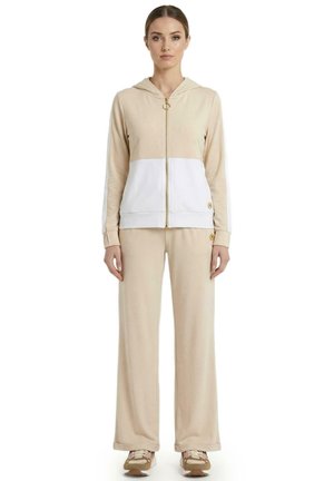 Une femme porte une veste beige à capuche et à fermeture éclair, avec un empiècement blanc dans le bas, assortie à un pantalon à jambes larges et à des baskets beiges.