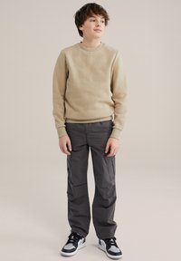 Beiger strukturierter Pullover mit geometrischem Muster, kombiniert mit dunkelgrauen Cargo-Hosen und schwarz-weißen Sneakers. Lässiges Outfit für Jugendliche.