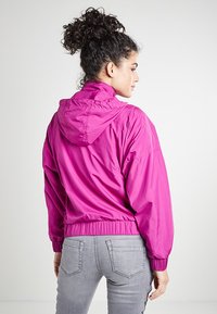 Lett rosa hettejakke med elastiske mansjetter og hem, med en glatt overflate og rynkedetalj bak.