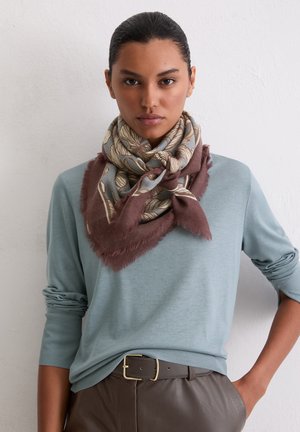 Frau mit nach hinten gekämmtem Haar, die ein blaues Langarmshirt, einen gemusterten Schal mit braunen Fransen und braune Lederhosen mit Gürtel trägt.