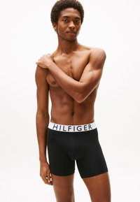 Boxer briefs negros con una cintura de logo "HILFIGER" en blanco, con una textura suave y un diseño ajustado, usados por un modelo con un fondo neutro.
