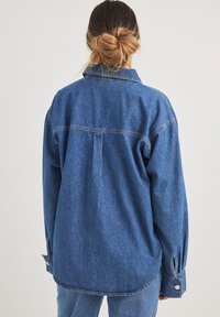 Übergröße Denim-Hemd in Mittelblau, mit einer Rückenfalte und knöpfbaren Manschetten. Weiche Textur mit sichtbaren Nähdetails.