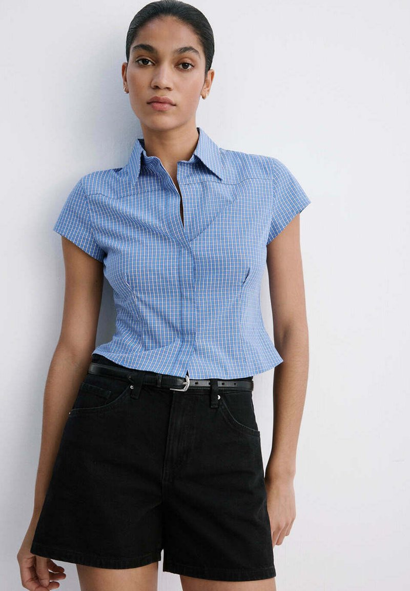 Jeune femme portant une chemise à carreaux bleue à manches courtes et un short noir taille haute, debout contre un mur blanc.