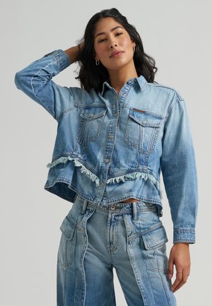 Frau trägt eine hellblaue, lässig abgenutzte Jeansjacke und passende hoch taillierte Jeans mit aufgesetzten Taschen, posiert mit der Hand im Haar.