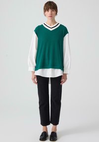 Canottiera a coste verde acqua con scollatura a V sopra una camicia bianca a maniche lunghe, abbinata a pantaloni neri a vita alta e mocassini neri. Design semplice, alto contrasto.