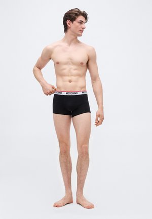 Άνδρας χωρίς πουκάμισο με μαύρα μποξεράκια Moschino τραβώντας τη μέση τους, όρθιος ξυπόλητος μπροστά σε απλό ανοιχτό φόντο, κοιτάζοντας προς τα πλάγια.