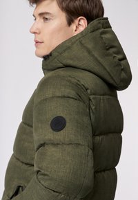 Grøn pufferjakke med en tekstureret overflade, der har en stor hætte og et sort rundt logo-patch på ærmet. Lynlåslommer synlige.