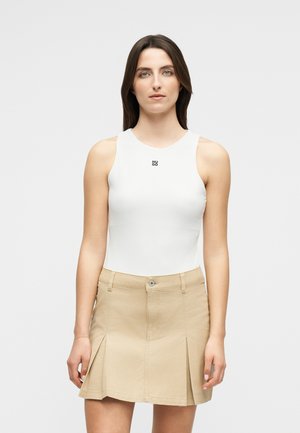 Γυναίκα με καστανά ίσια μαλλιά, φορώντας άσπρο αμάνικο τοπ με το λογότυπο "HUGO" και μπεζ πλισέ μίνι φούστα, στέκεται μπροστά σε απλό φόντο.