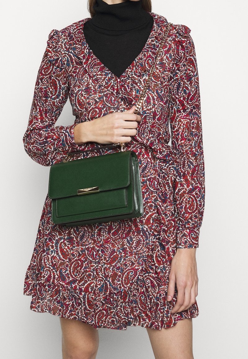 Femme portant une robe rouge à motif paisley sur un col roulé noir, tenant un sac à main en cuir vert foncé avec une bandoulière chaîne dorée.