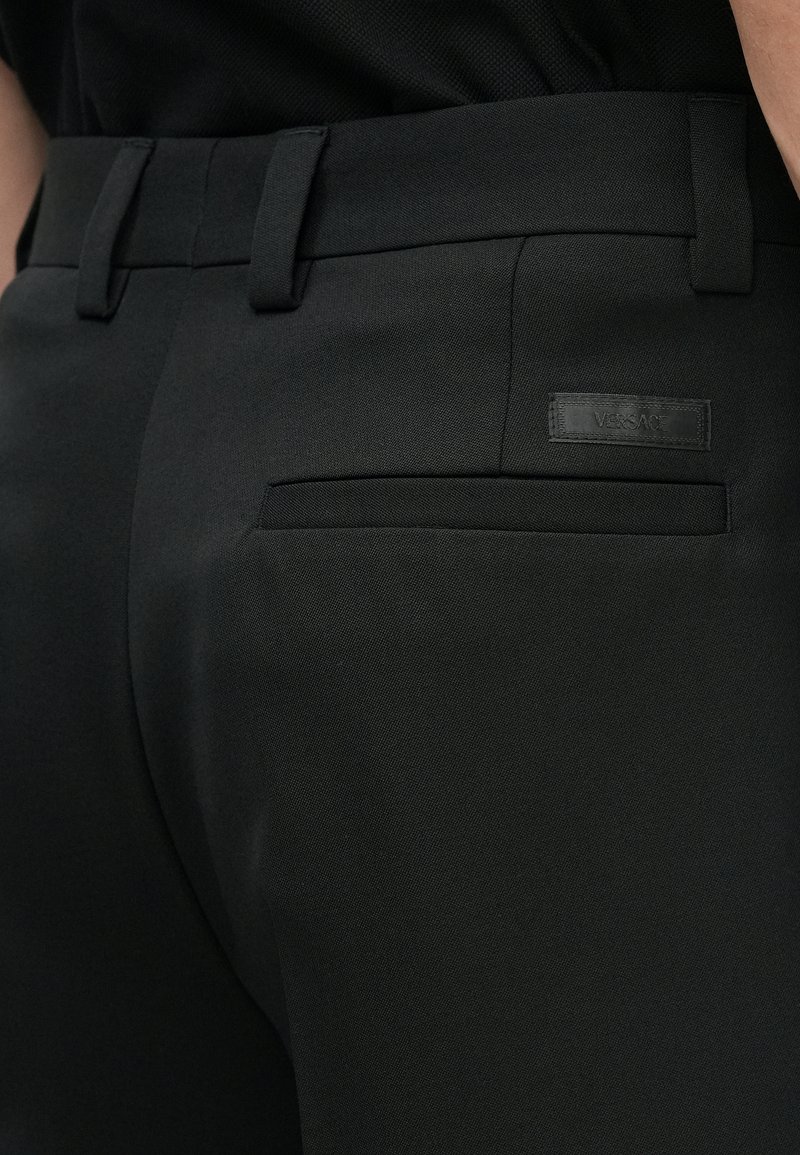 Close-up of black tailored pants with belt loops, a back welt pocket, and a small black Versace logo patch above the pocket.