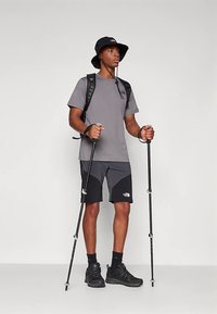 Pilka techninė marškinėliai, juodos žygio šortai su logotipu, juodi žygio batai, laikantys žygio lazdas, juoda kibiro formos kepurė ir kuprinė.