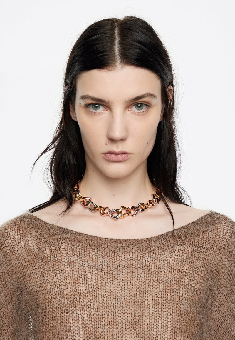 Collar de cadena metálica con eslabones geométricos entrelazados en tonos dorado, plateado y rosado, usado sobre un suéter marrón de punto suelto.