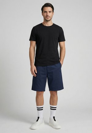 Mann steht mit Händen in den Taschen, trägt ein schwarzes T-Shirt, marineblaue Shorts, weiße Socken mit schwarzen Streifen und weiße Slip-On-Schuhe.