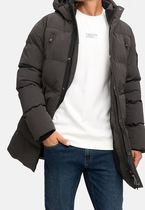 Mann trägt eine dunkelgraue gesteppte Jacke über einem weißen Sweatshirt mit dem Text "Wear the Code" und eine blaue Jeans, die Hand in der Tasche.