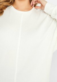 Bílý žebrovaný top s dlouhými rukávy, kulatým výstřihem a texturovaným materiálem. Mírně volný střih s viditelnými švy.