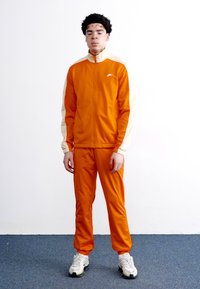 Πορτοκαλί αθλητική φόρμα με τζάκετ που κλείνει με φερμουάρ και αντίστοιχους παντελόνες. Το τζάκετ έχει κρεμ λεπτομέρειες στα μανίκια. Φοριέται με λευκά αθλητικά παπούτσια.