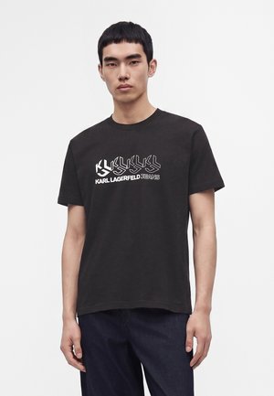 Νέος άνδρας που φοράει μαύρο t-shirt Karl Lagerfeld Jeans με λευκή εκτύπωση λογοτύπου, στέκεται μπροστά σε απλό ανοιχτό φόντο.