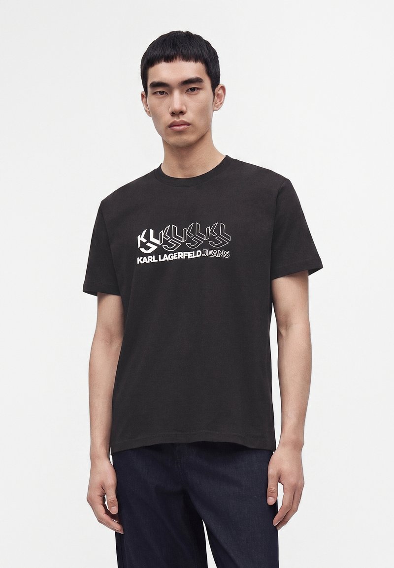 Νέος άνδρας που φοράει μαύρο t-shirt Karl Lagerfeld Jeans με λευκή εκτύπωση λογοτύπου, στέκεται μπροστά σε απλό ανοιχτό φόντο.