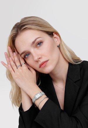 Blondynka o niebieskich oczach w czarnej kurtce, opierająca ręce przy twarzy, nosząca srebrne pierścionki, bransoletkę, zegarek i małe kolczyki kółka.