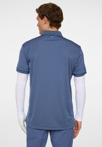 Blå kortärmad polo med krage och fukttransporterande tyg, visad bakifrån, med en logotyp vid nacken.