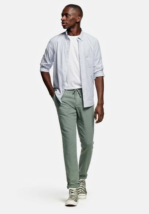 Man walking, wearing light blue striped button-up shirt over white t-shirt, green drawstring pants, and green high-top sneakers.