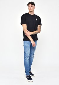 Musta puuvillapaita, jossa on pieni logopatch, yhdistettynä vaaleansinisiin farkkuihin ja tummansinisiin lenkkareihin. Malli seisoo kädet ristissä.