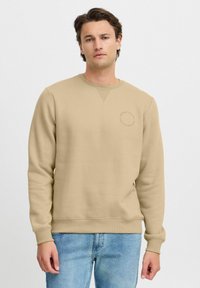 Beige sweatshirt lavet af en blanding af bomuld, med rund hals, ribbede manchetter og et lille cirkellogo på brystet. Båret med blå jeans.