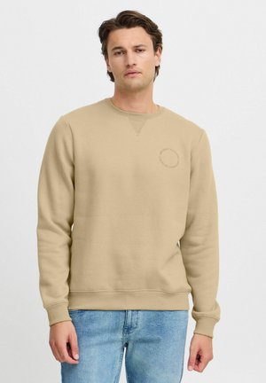 Beige sweatshirt made of cotton-blend fabric, featuring a crew neck, ribbed cuffs, and a small circle logo on the chest. Worn with blue jeans.