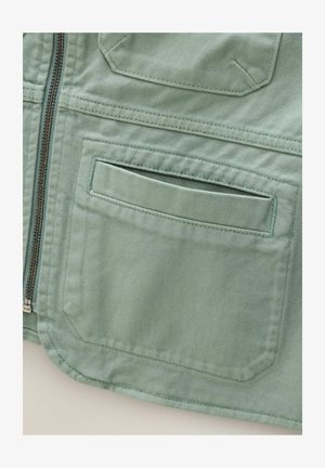 Gaiši zaļa ar rāvējslēdzēju teļa jaka, kurai ir diagonāla faktūra, taisnstūra krūšu kabata un kārtīgas šuves gar malām.