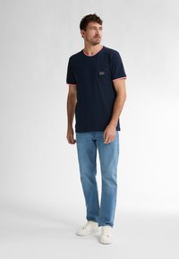 Marinblå t-shirt med kort ärm och röd kant på krage och ärmar, med en bröstficka med en logotagg, sammansatt med ljusblå jeans och vita skor.