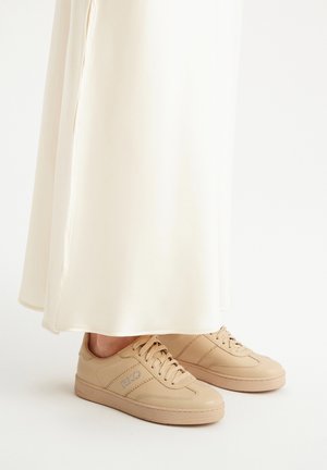 Sneakers in pelle beige con punta rotonda, laccatura frontale e dettaglio del logo sul lato, abbinati a un vestito fluente di colore crema.