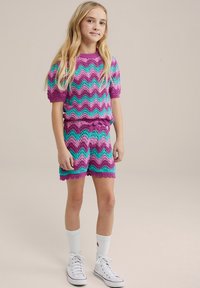 Pletený dvojdielny outfit so vzorom v fialovej, tyrkysovej a ružovej farbe v tvare zúbkovej stopy. Top s krátkym rukávom a zodpovedajúca sukňa so zúbkovanými okrajmi. Biely tenisky.