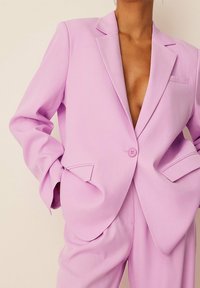 Osoba ubrana w jasnofioletowy, dopasowany garnitur z marynarką na jeden guzik, z rękoma w kieszeniach i bez widocznej koszuli pod spodem.