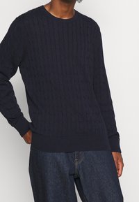 Pullover blu navy con lavorazione a trecce, scollo rotondo, polsini e orlo a coste, con un motivo testurizzato in tutto il capo per maggior dettaglio.