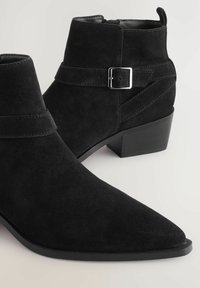 Botas de tobillo de ante negro con punta afilada y un pequeño tacón cuadrado. Presenta una correa con una hebilla plateada y acentos texturizados.
