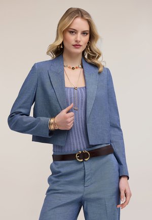 Blonde vrouw in een blauwe cropped blazer, bijpassende broek, gestreepte paarse top, bruine riem, gouden sieraden, poseert met één hand dichtbij haar borst.