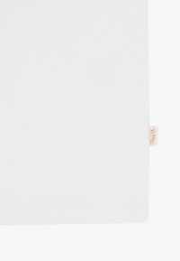 Κοντινό πλάνο λευκού ριμπ ύφανσης με ραμμένη στριφώματα και μια μικρή μπεζ ετικέτα με την ένδειξη "BLKVS" στη δεξιά άκρη.