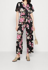 Mono negro de flores con mangas cortas, adornado con flores rosas y blancas, cintura ceñida y pantalones de pierna ancha. Combinado con sandalias de tacón cuadrado en color beige.