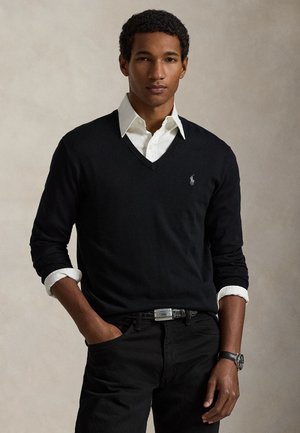 Homme aux cheveux courts et bouclés porte une chemise blanche à col sous un pull noir à col en V et un pantalon noir, debout avec une main dans la poche.