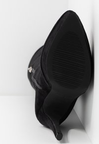 Botte à talon haut en daim noir à bout pointu vue par la semelle, appuyée contre une surface blanche.