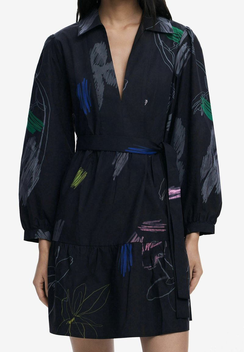 Femme portant une robe sombre avec de longues manches bouffantes, des motifs floraux et abstraits colorés, un col, un décolleté en V profond et une ceinture nouée à la taille.