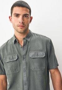 Short-sleeve, collared shirt in muted green fabric, featuring two chest pockets and buttoned front with grey accents and a smooth texture.
