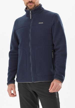 Homme portant une veste polaire bleu marine à fermeture éclair intégrale avec poches latérales zippées et un pantalon de plein air noir.
