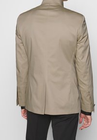 Beige skräddarsydd kavaj med hög krage, enkel ventilering bak och textilier med struktur. Har en avslappnad passform och minimalistisk design.