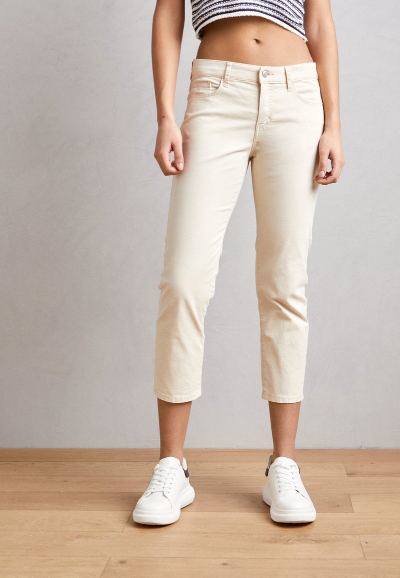 Beige cropped broek met een slanke pasvorm, gemaakt van een zacht materiaal, gecombineerd met witte sneakers met een zwarte accenten op de hiel.