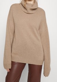 Beige oversized trui met een geribbelde col, textuur breiwerk en lange mouwen; gecombineerd met bruine faux leren broek.